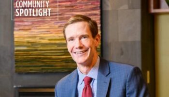 A Smiling Man In A Suit And Red Tie Poses Indoors In Front Of A Colorful, Abstract Wall Artwork. The Words Community Spotlight Appear In The Upper Left Corner Of The Image.