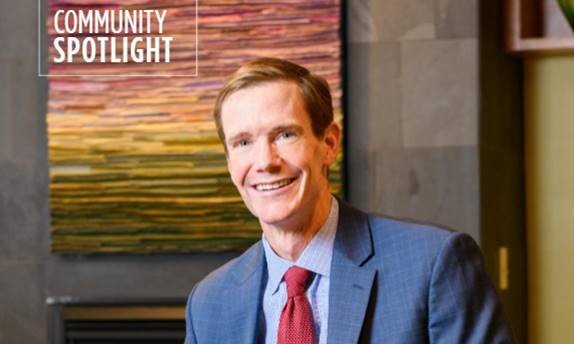 A smiling man in a suit and red tie poses indoors in front of a colorful, abstract wall artwork. The words Community Spotlight appear in the upper left corner of the image.