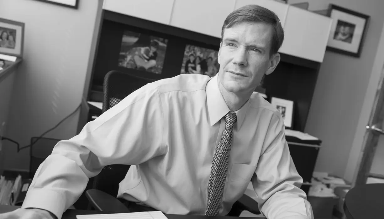 A man in a shirt and tie sits at a desk in an office, looking thoughtfully to the side. There are shelves, framed photos, and office supplies in the background. The image is in black and white.