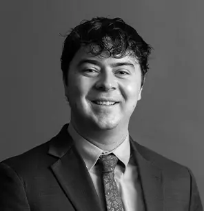 A man with dark, wavy hair wearing a suit, dress shirt, and patterned tie smiles at the camera against a plain, gray background. The image is in black and white.