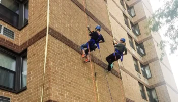 Two People Wearing Helmets And Harnesses Rappel Down The Side Of A Tall Brick Building, Using Ropes For Support. They Are Facing The Wall And Appear To Be Participating In An Outdoor Climbing Activity.