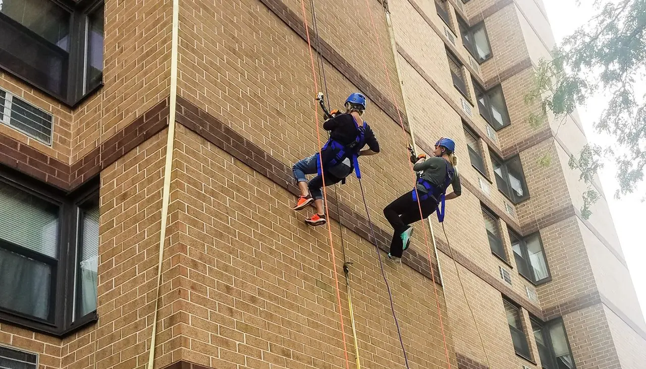 Two people wearing helmets and harnesses rappel down the side of a tall brick building, using ropes for support. They are facing the wall and appear to be participating in an outdoor climbing activity.