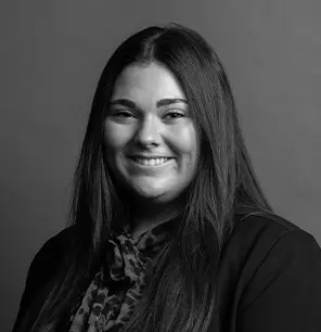 Black and white portrait of a woman with long straight hair, smiling, wearing a dark blazer and a patterned blouse, posed in front of a plain background.