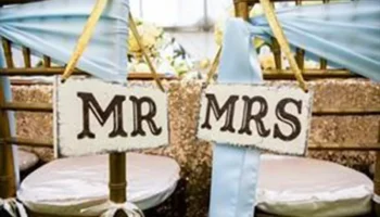 Two Chairs Decorated With Blue Fabric And Gold Ribbons, Each With A Sign On The Back Reading “MR” And “MRS,” Suggesting A Wedding Or A Celebration For A Married Couple.