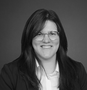 A person with long dark hair and glasses, wearing a suit jacket over a button-up shirt, smiles at the camera against a plain dark background. The image is in black and white.