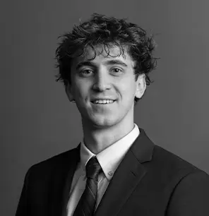 A young man with curly hair, wearing a suit, dress shirt, and tie, smiles at the camera against a plain, dark background. The image is in black and white.