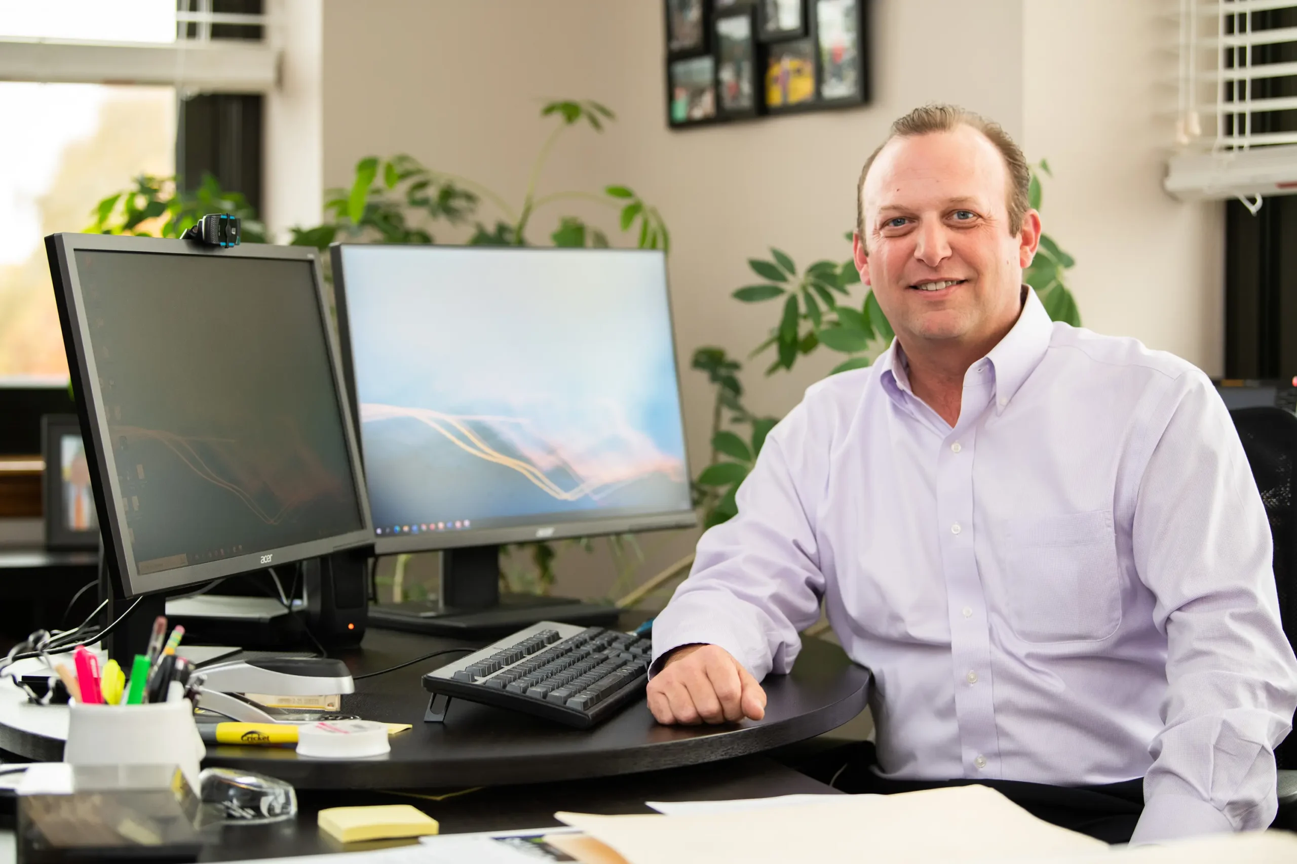 A man in a light purple shirt sits at a desk in an office, smiling at the camera. There are dual monitors, office supplies, plants, and papers visible on the desk and in the background.