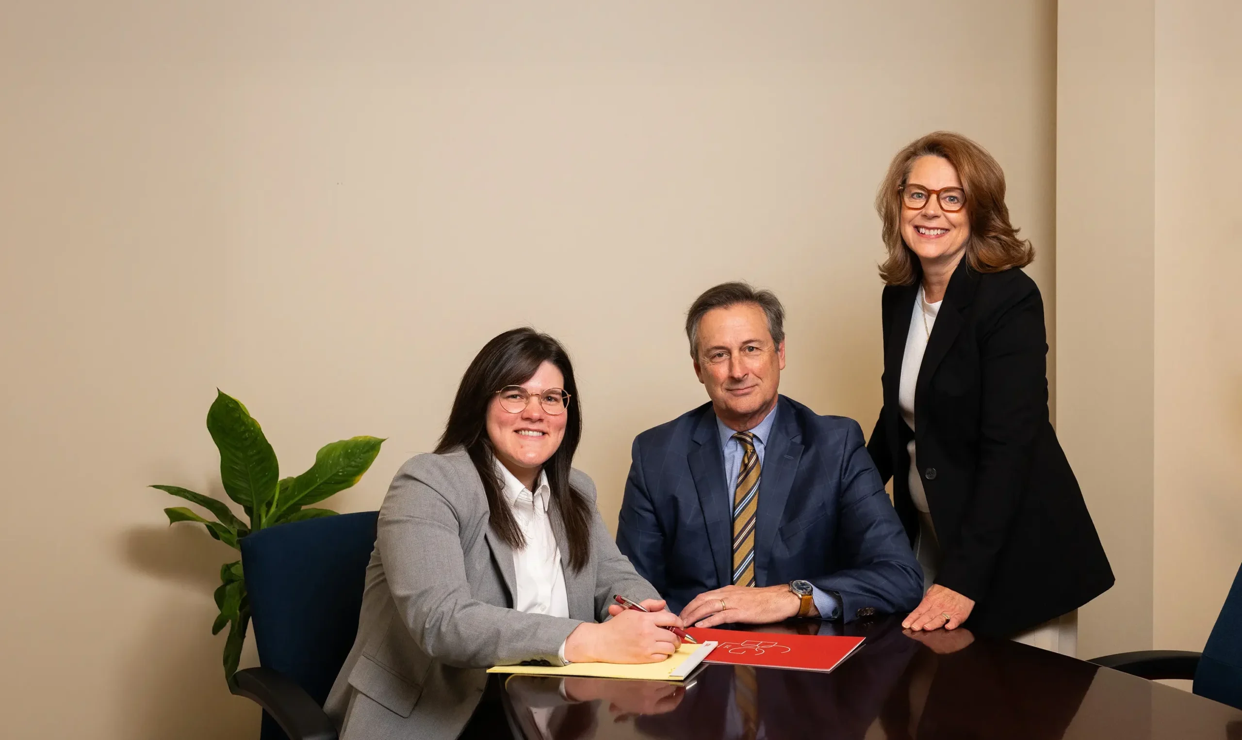 Three professionally dressed people, two women and one man, sit and stand around a conference table with documents. One woman is writing, the man sits at the center, and the other woman stands, all smiling at the camera. A plant is in the background.