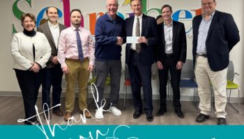 Six Adults, Five Men And One Woman, Stand Smiling In A Row Indoors. Two Men Shake Hands While Holding A Check. The Background Features Colorful Schreiber Letters And A Blue Banner That Says Thank You BG LLC.