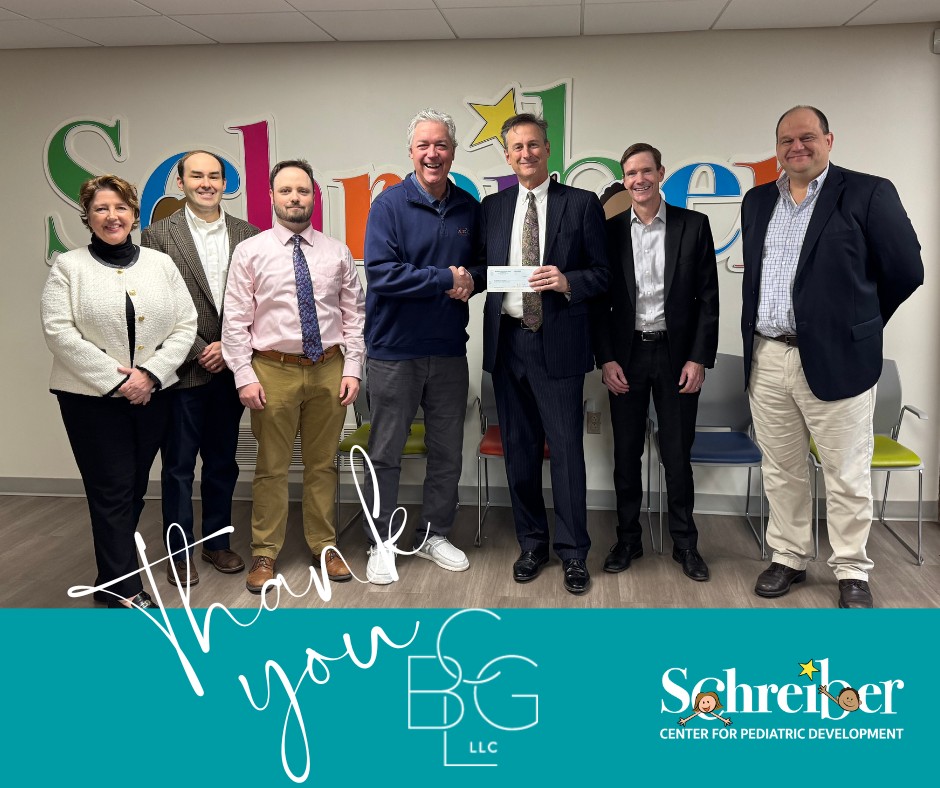 Six adults, five men and one woman, stand smiling in a row indoors. Two men shake hands while holding a check. The background features colorful Schreiber letters and a blue banner that says Thank you BG LLC.