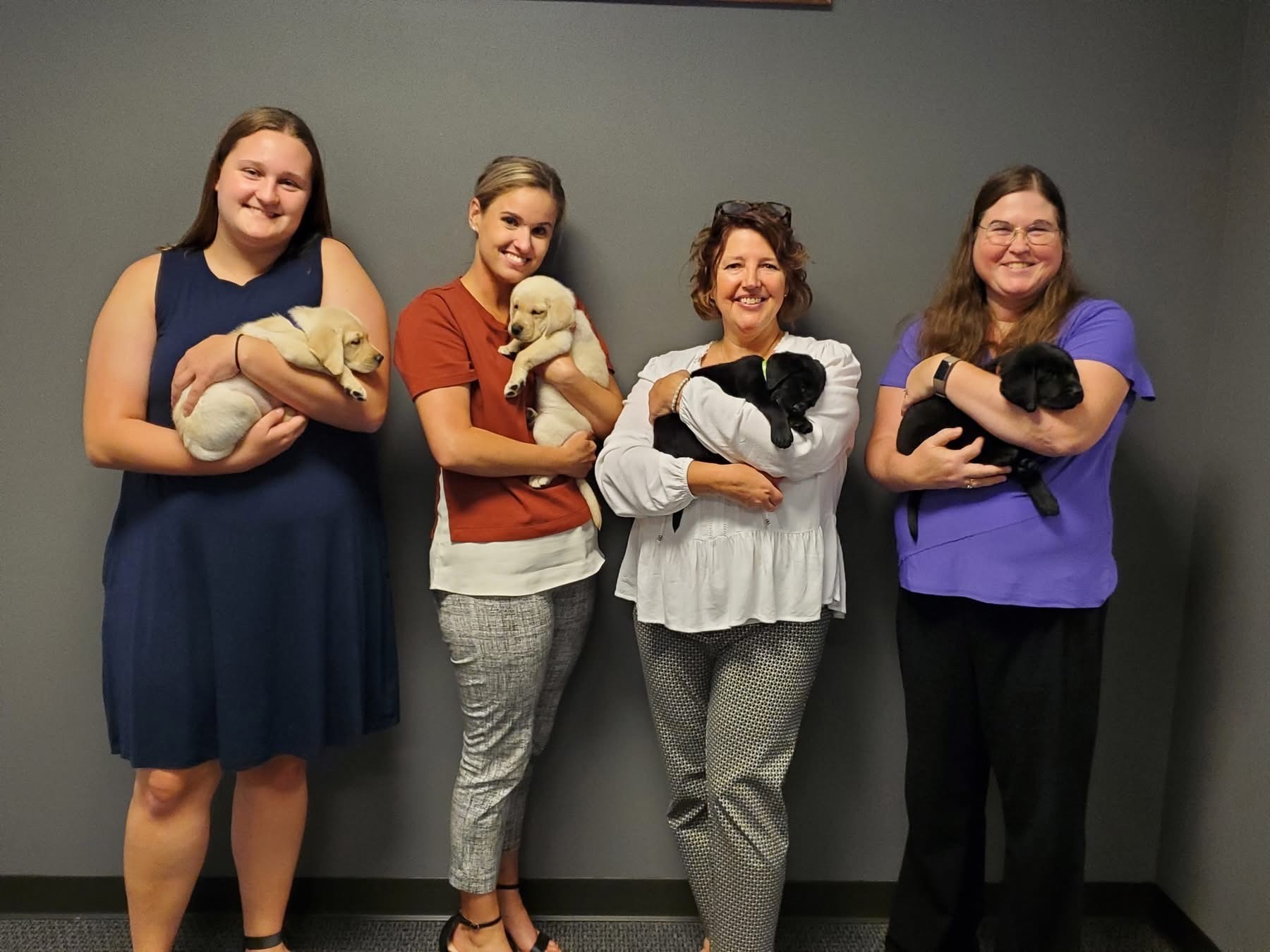 Four women stand side by side indoors, each smiling and holding a puppy; two women hold yellow puppies and two hold black puppies, all against a gray wall background.