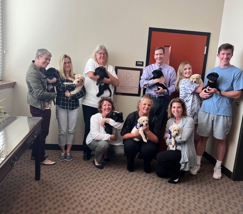 A group of ten smiling people, some standing and some kneeling, each holding a puppy of various colors, pose indoors in an office with beige walls and brown carpet. A sign is mounted on the wall behind them.