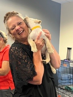 A woman smiles and laughs as a small puppy with a yellow collar licks her face. She is holding the puppy close in her arms, and another person stands in the background.