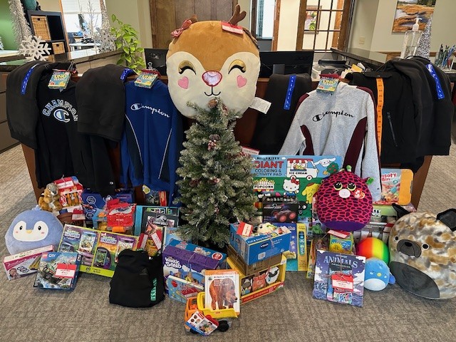 A small decorated Christmas tree is surrounded by various toys, stuffed animals, board games, books, and clothing, including Champion sweatshirts. A large reindeer plush sits behind the tree. The scene is indoors.