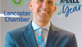 A Smiling, Bald Man In A Blue Suit And Plaid Tie Stands With Arms Crossed In Front Of The Lancaster Chamber Logo And The Text Small Businessperson Of The Year.