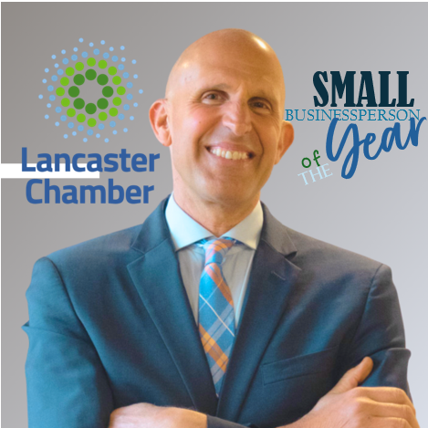 A smiling, bald man in a blue suit and plaid tie stands with arms crossed in front of the Lancaster Chamber logo and the text Small Businessperson of the Year.
