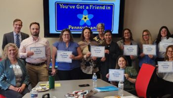 A Group Of People Pose In A Conference Room Holding Signs That Say Youve Got A Friend In Pennsylvania. There Are Snacks And Drinks On The Table, And A Similar Message Is Displayed On The Screen Behind Them.