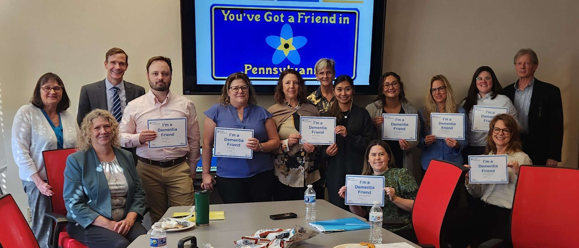 A group of people pose in a conference room holding signs that say Youve Got a Friend in Pennsylvania. There are snacks and drinks on the table, and a similar message is displayed on the screen behind them.